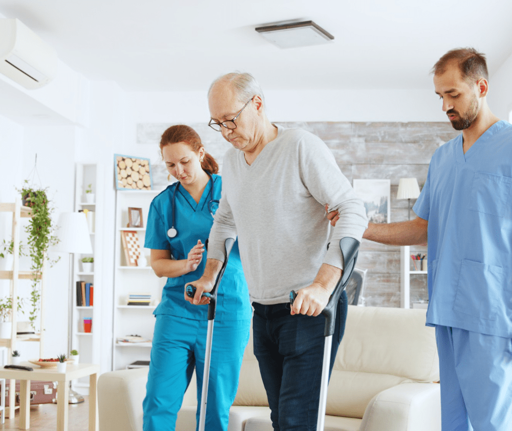 Team of nurses or social workers helping an old disabled man to walk with his crutches out of the nursing home room.